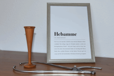 ein hölzernes Stethoskop steht neben einem Bilderrahmen. Darin ist ein Bild mit dem Titel "Hebamme". Im Vordergrund liegt ein Beckenzirkel.