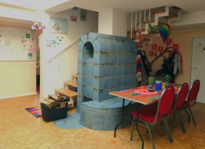 zentral ist ein großer blauer Kachelofen mit Ofenbank zu sehen. Davor laden ein Tisch und Stühle zum gemütlichen Beisammensein ein.
